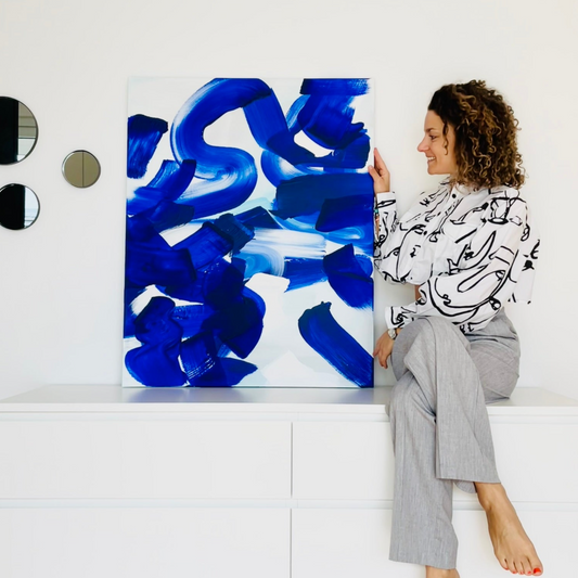 FLOR. sitzend auf einer weißen Kommode mit ihrem abstrakten originalen Gemälde das vor blauen Farben funkelt. 